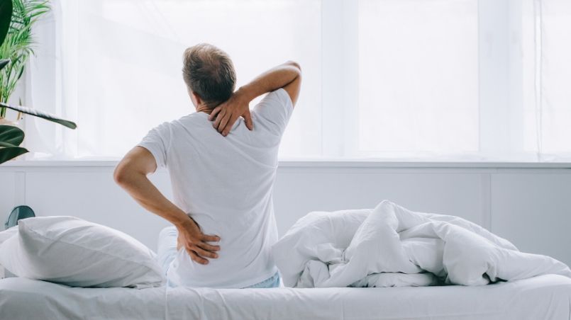 Man sitting on edge of bed with lower back pain and should neck pain