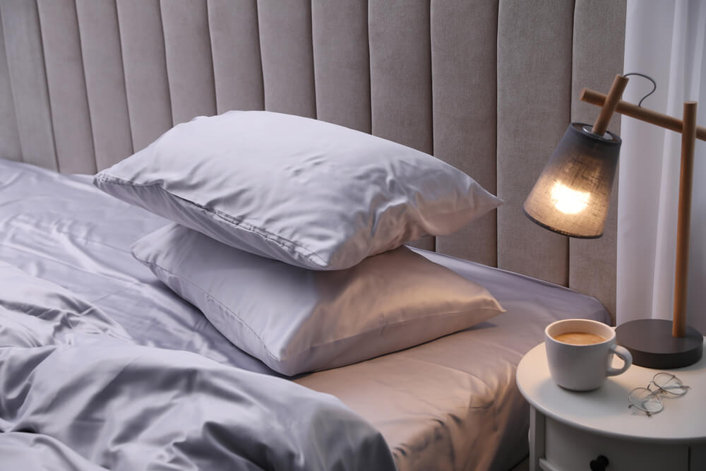 A bed with beautiful light grey pillows in silk bedding and pillowcases and a grey headboard with side lamp and a cup of coffee