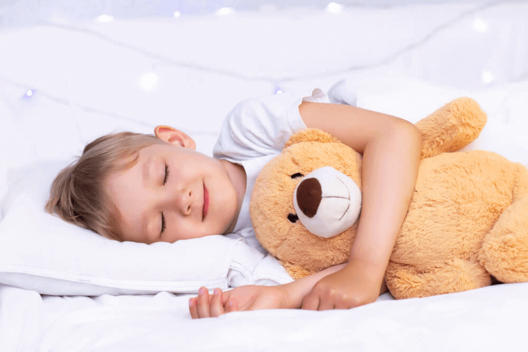 A toddler sleeping in bed on a pillow and duvet cover with a soft toy