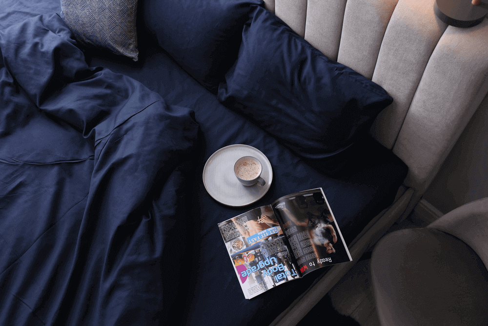 Stylish bed, silky navy bed sheets and linen, coffee, magazine and grey headboard