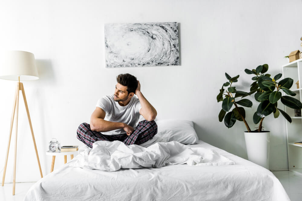 A young man sitting in bed in a white themed bedroom with green plants and beautiful artwall and simple clothes rail.