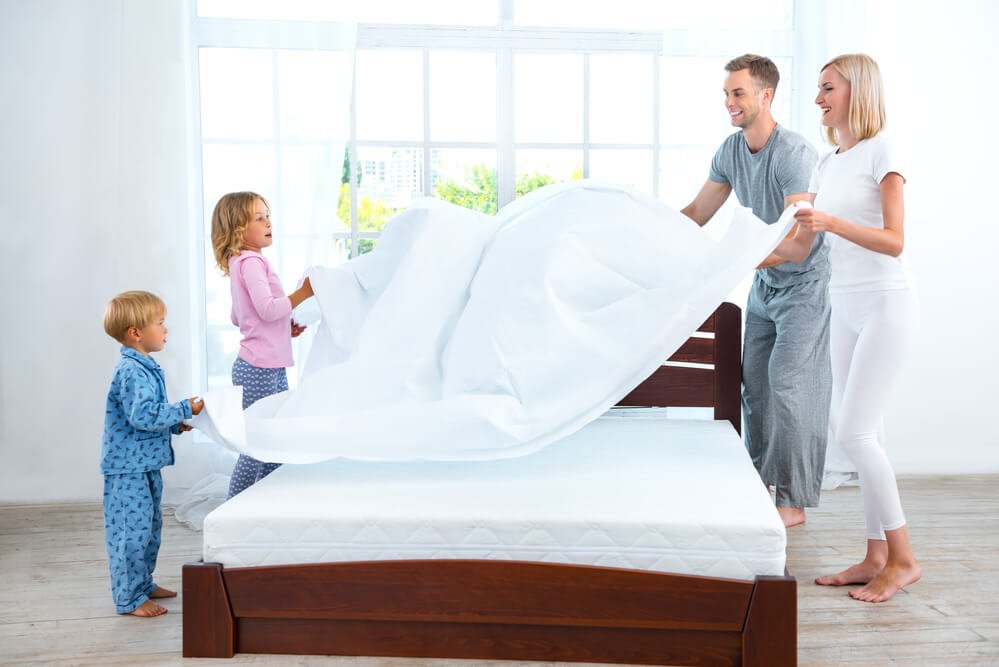 A family of four helping making a bed and changing bed sheets on an encased mattress.