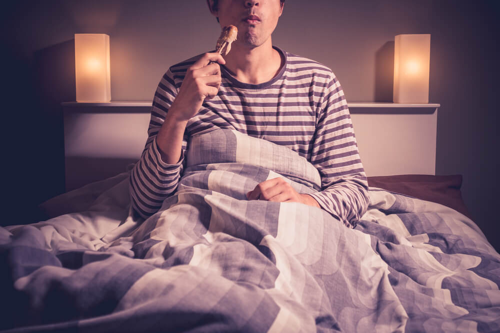 A young male eating a chicken drumstick in bed at midnight