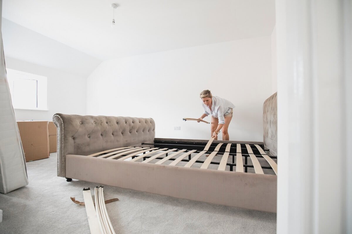 A woman putting together her new bed . She is placing wooden slats on the bed frame